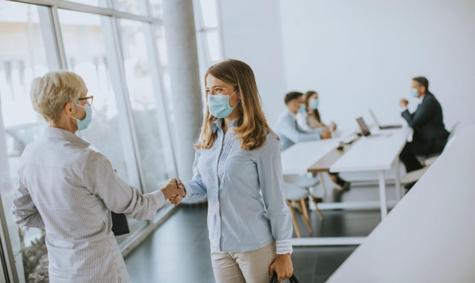 Two businesswomen, mature and young one, handshaking in the office and wearing mask as a virus protection