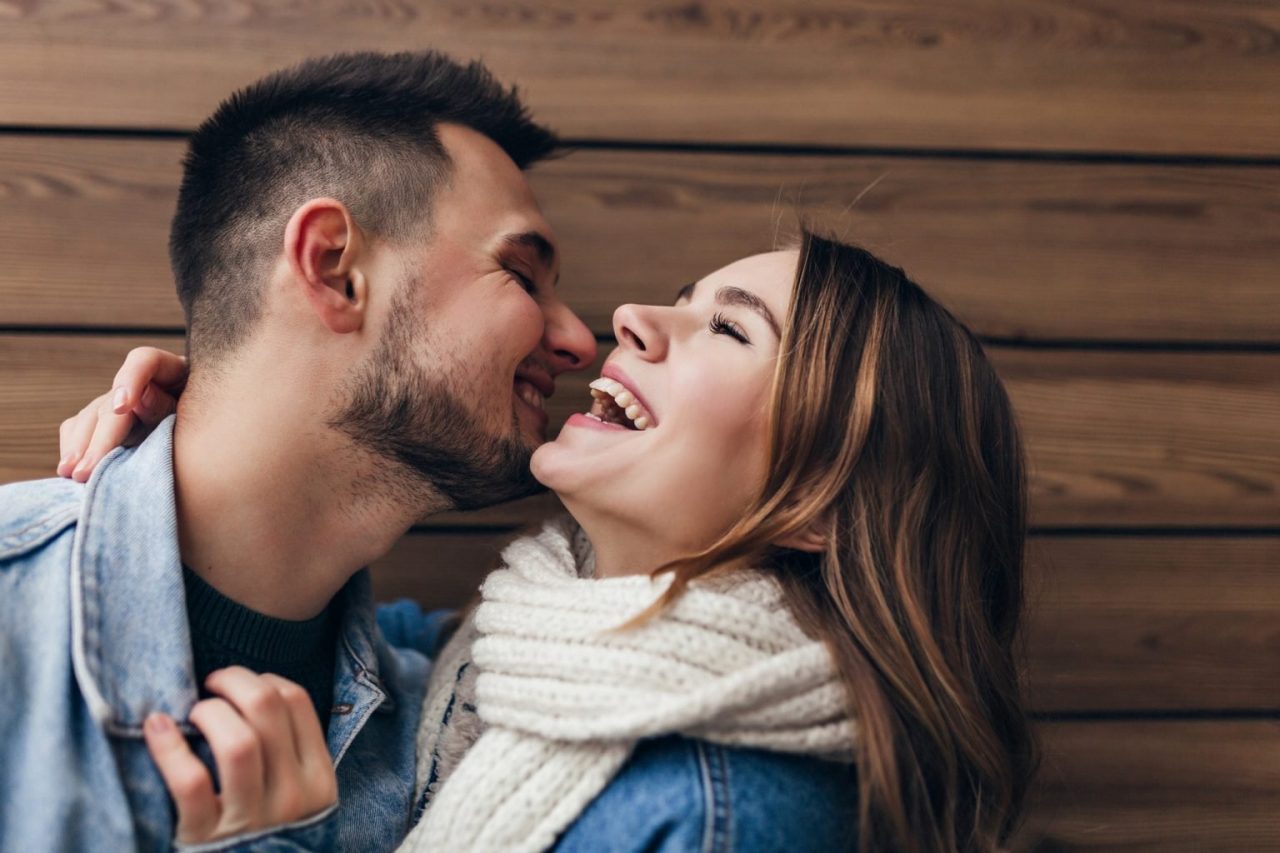 laughing-brunette-man-expressing-love-while-posing-with-girlfriend-blissful-caucasian-couple-kissing-wooden-wall-1536x1024