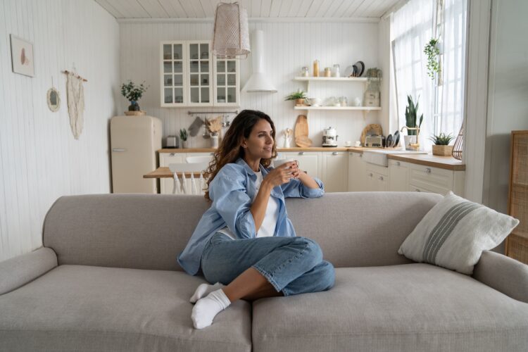 Independent woman dreaming, holding cup of coffee sits on sofa in spacious house enjoying loneliness